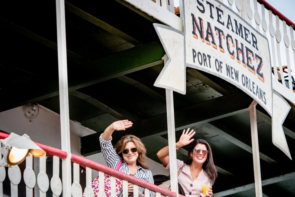 Two women wave from the back of the Natchez. A sign reads: "Steamboat Natchez, Port of new Orleans" in a typeface reminiscent of the Wild West