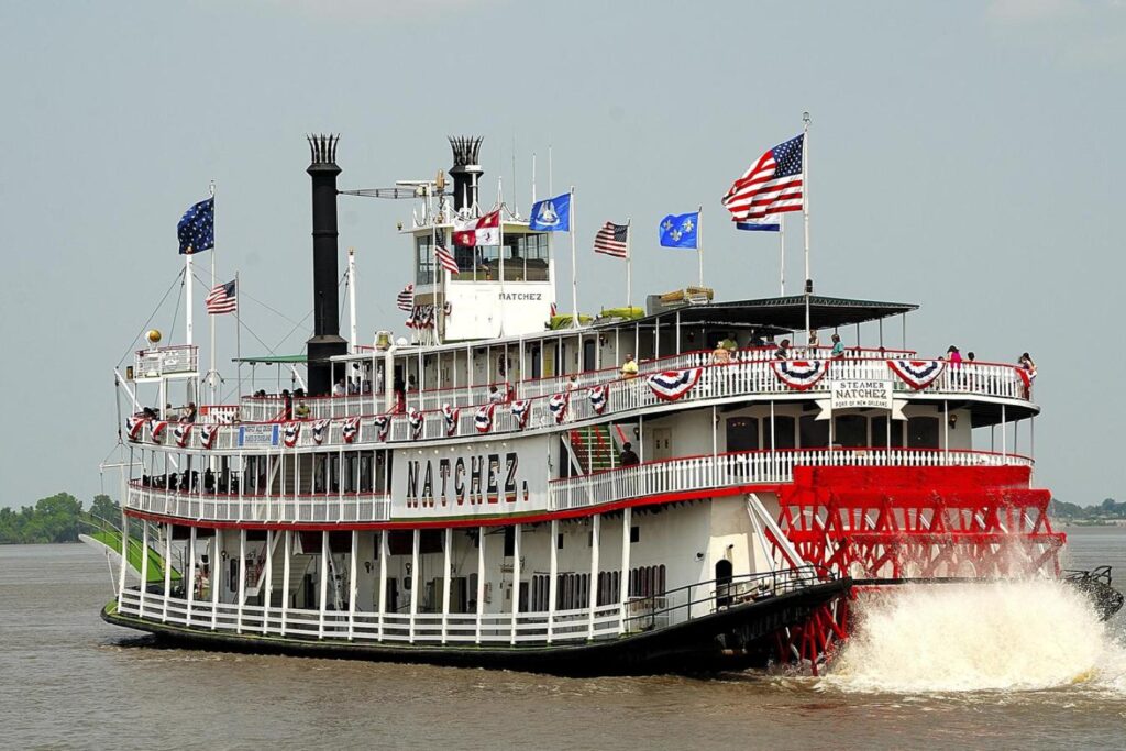 A wide shot of the Steamboat Natchez sailing down the Mississippi. Her red paddlewheel is churning the water behind her