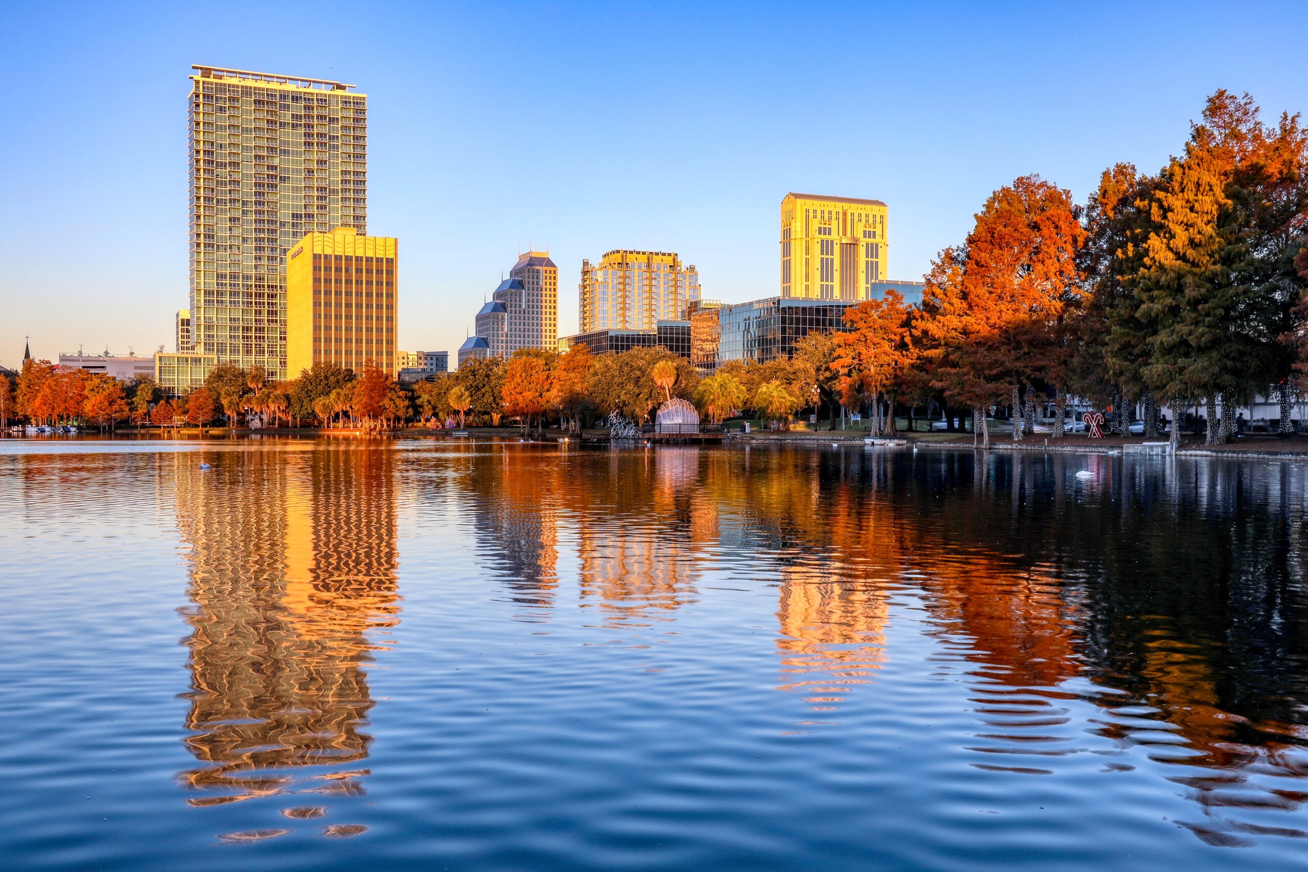 The sun rises over Lake Eola Park in Orlando, Florida. The still lake reflects trees and skyscrapers bathed in orange light