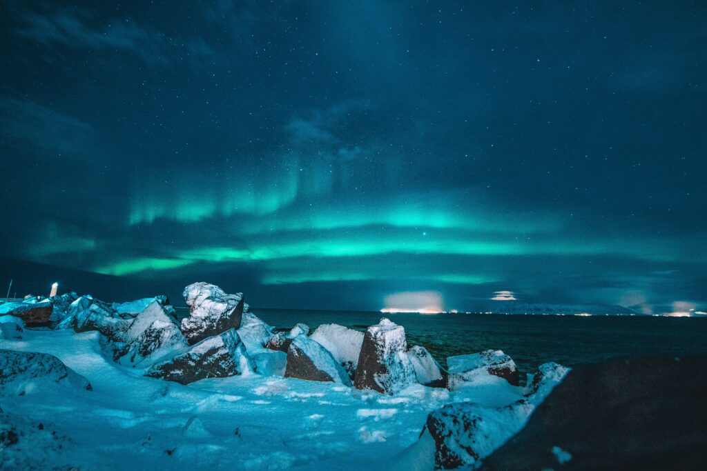 Bands of the Northern Lights can be seen stretched across the sky over the sea. Ice flows and rocks are in the foreground