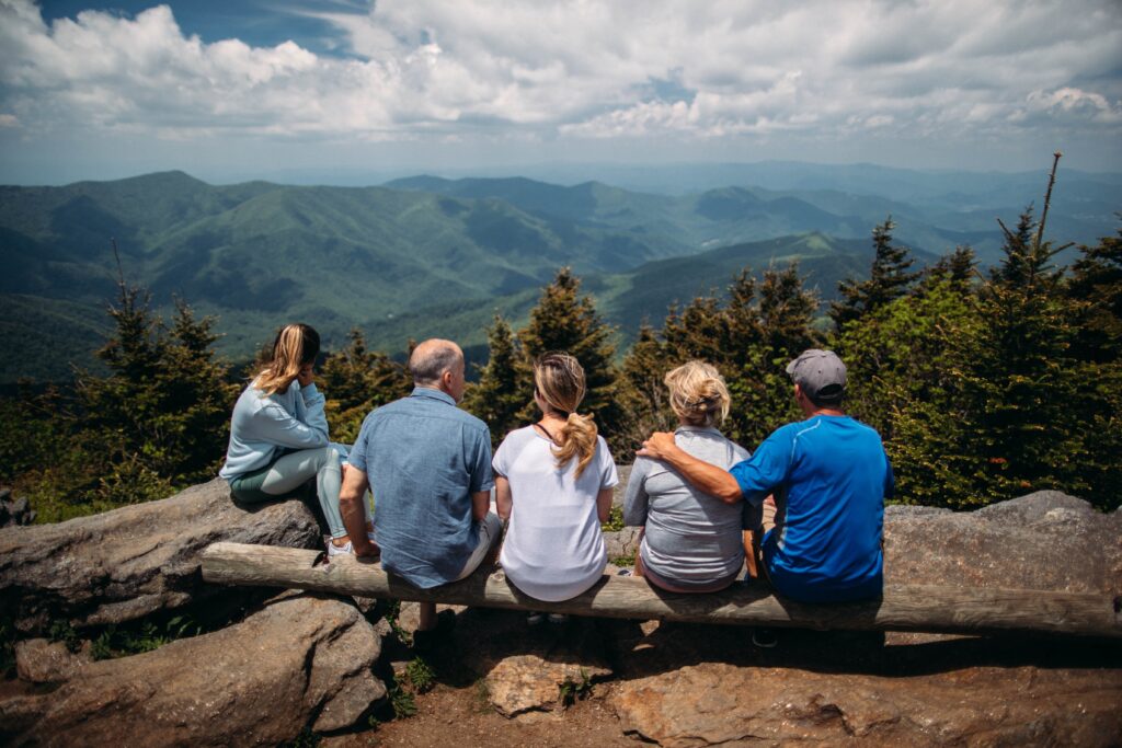 Three generations of a family sit on a log and look out over the Blue Ridge Mountains outside of Asheville. The father has his hand around the mother's shoulders
