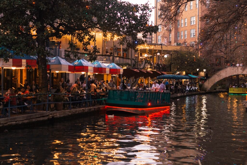 People sit beside the river under awnings bearing the Texan flag in the city of San Antonio