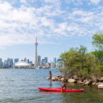 A person in a canoe launches from a bank on Lake Ontario with the Toronto Skyline in the background