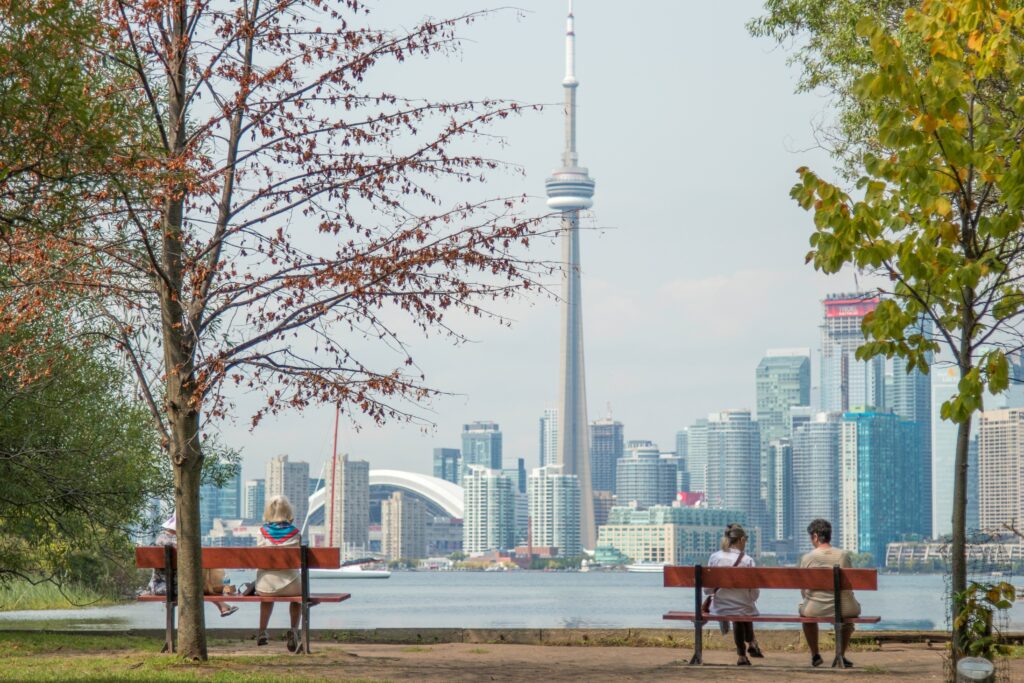 People sit on park benches on the edge of Lake Ontario. The distinctive shape of the CN Tower can be seen through the trees