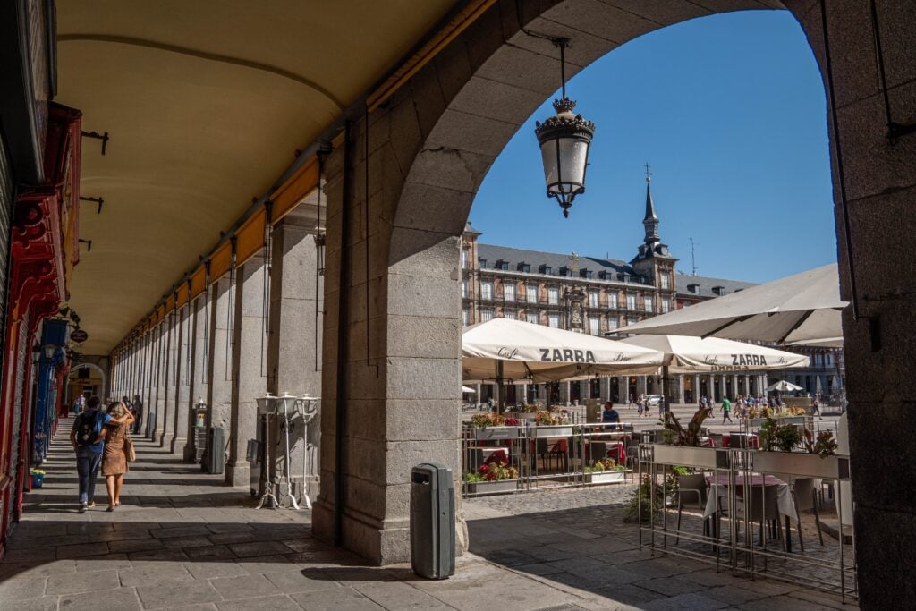 People walk under the arches of Plaza Mayor. The umbrellas of an outdoor restaurant can be seen and Casa de Panaderia is in the background