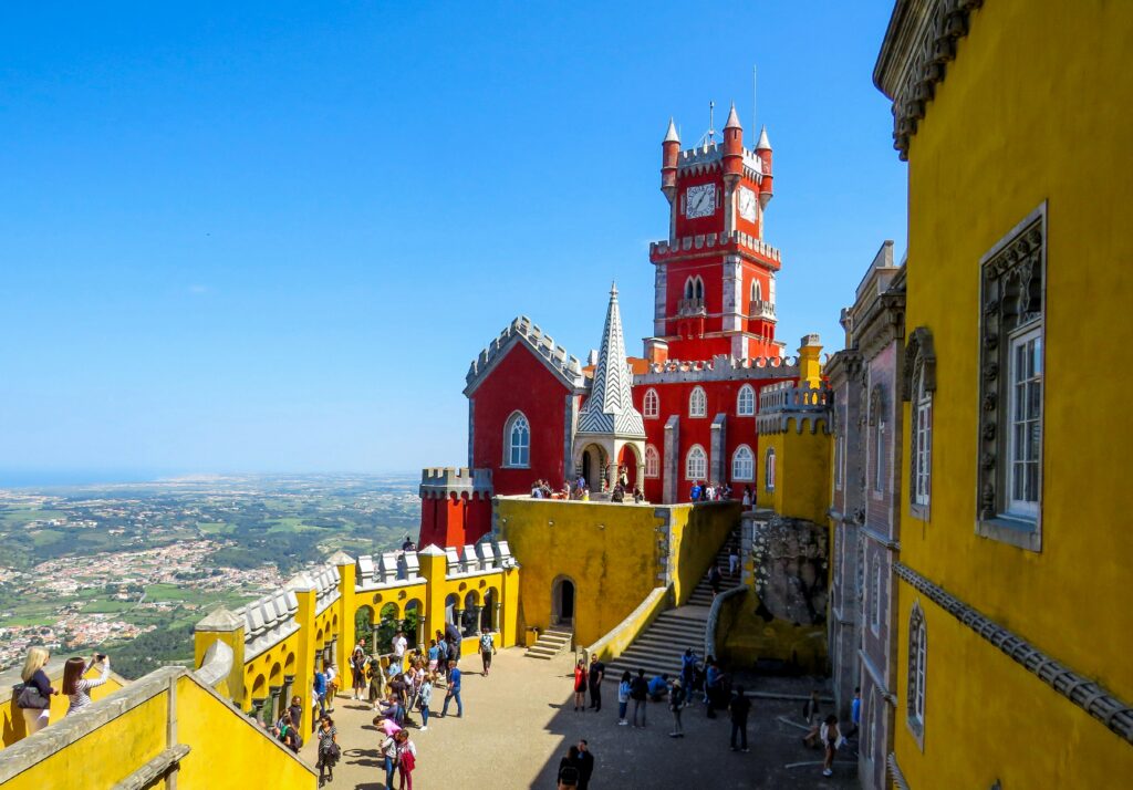 People walk through a yellow courtyard in Pena Palace with the red clock tower above them